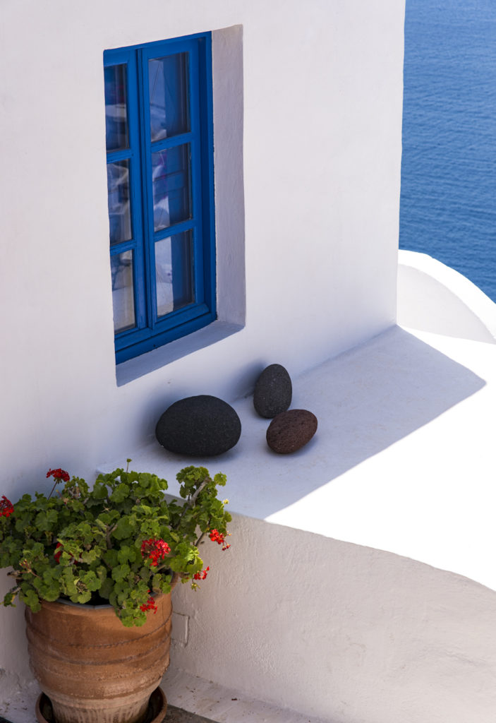 blaues Fenster eines weissen Hauses auf Santorini, davor schwarze Steine und rote Geranien