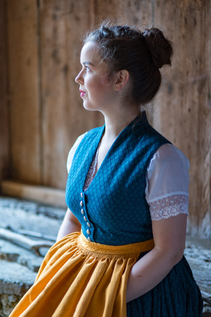 Portrait Fotografie, Available Light: blau-gelbes Dirndl im Halbportrait seitlich im Gegenlicht vor Holzwand