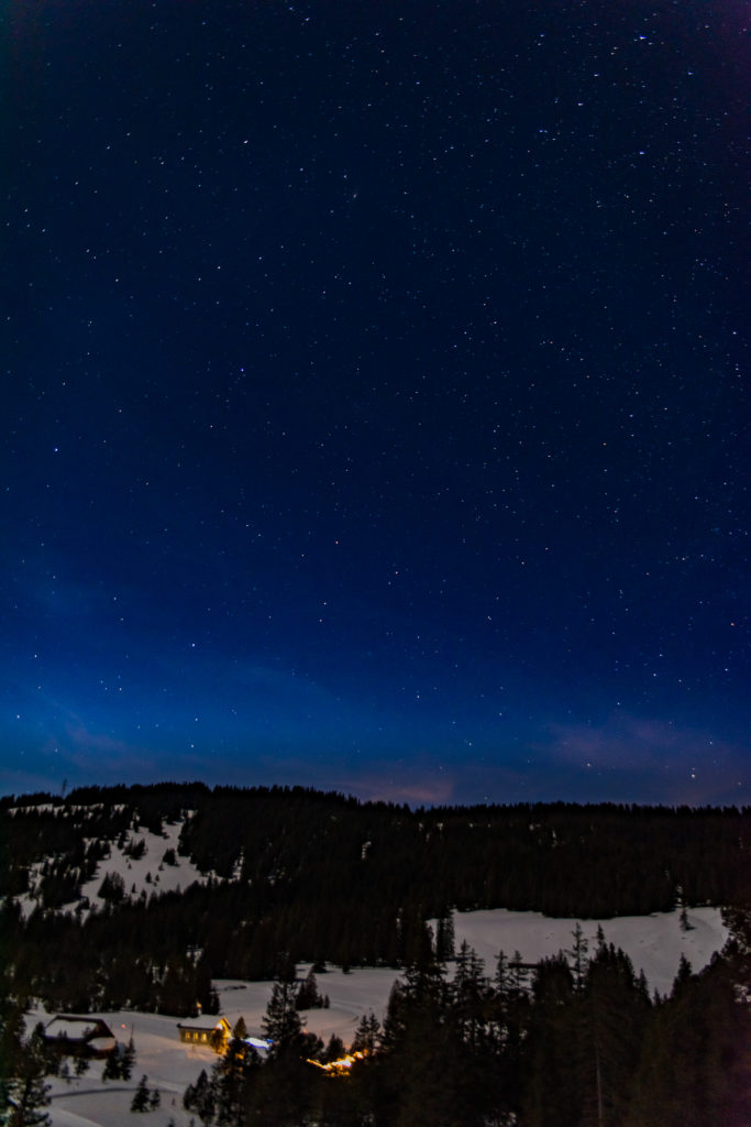 Winternacht mit klarem Sternenhimmel und beleuchteter Kapelle in einer Waldlichtung