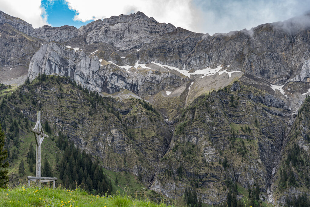 felsige Bergkette mit dunklen Quellwolken, im Vordergrund eine Bergwiese mit einem hölzernen Gipfelkreuz