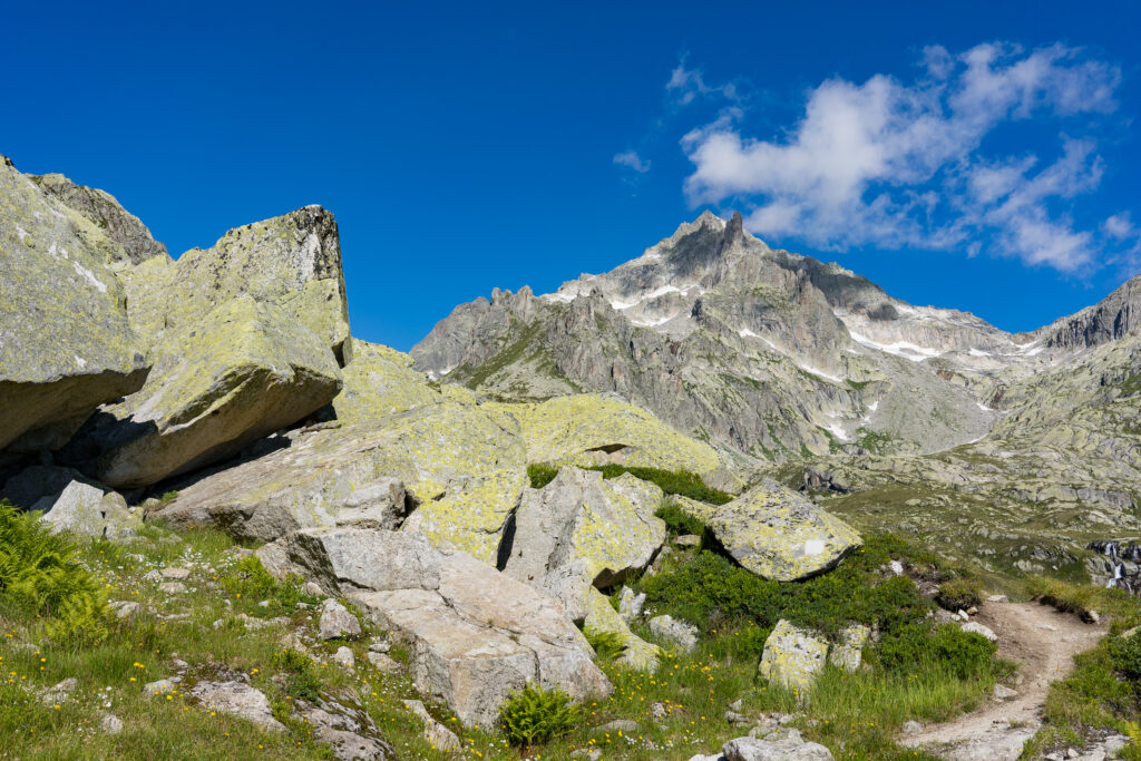 Wanderweg zwischen grossen Felsblöcken, im Hintergrund steil aufragende Felsgipfel