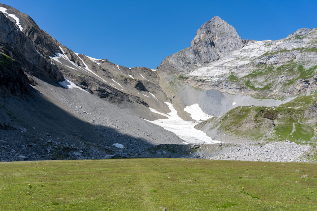 im Vordergrund ebene, grüne Gebirgswiese, im Hintergrund schroffe Felsabbrüche und steile Gipfel