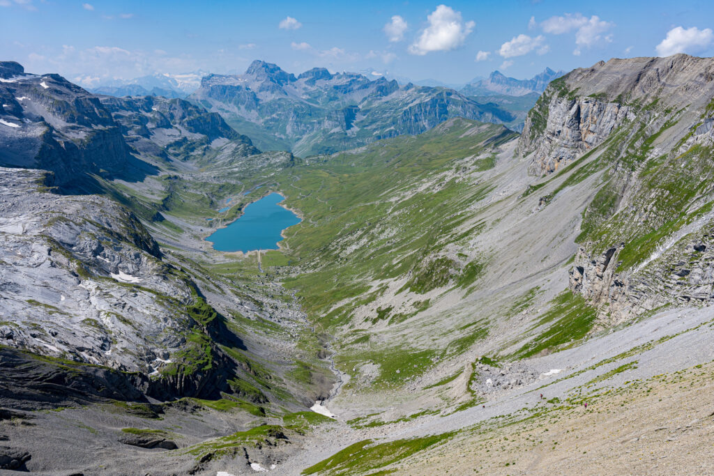 Ausblick vom Berggipfel auf ein Hochtal mit Bergsee, im Hintergrund Berge und Himmel mit Quellwolken