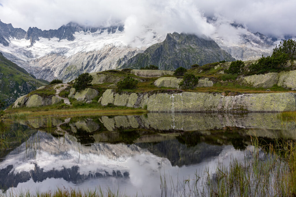 Bergsee mit spiegelnden in Wolken verhüllten Berggipfeln und Gletschern