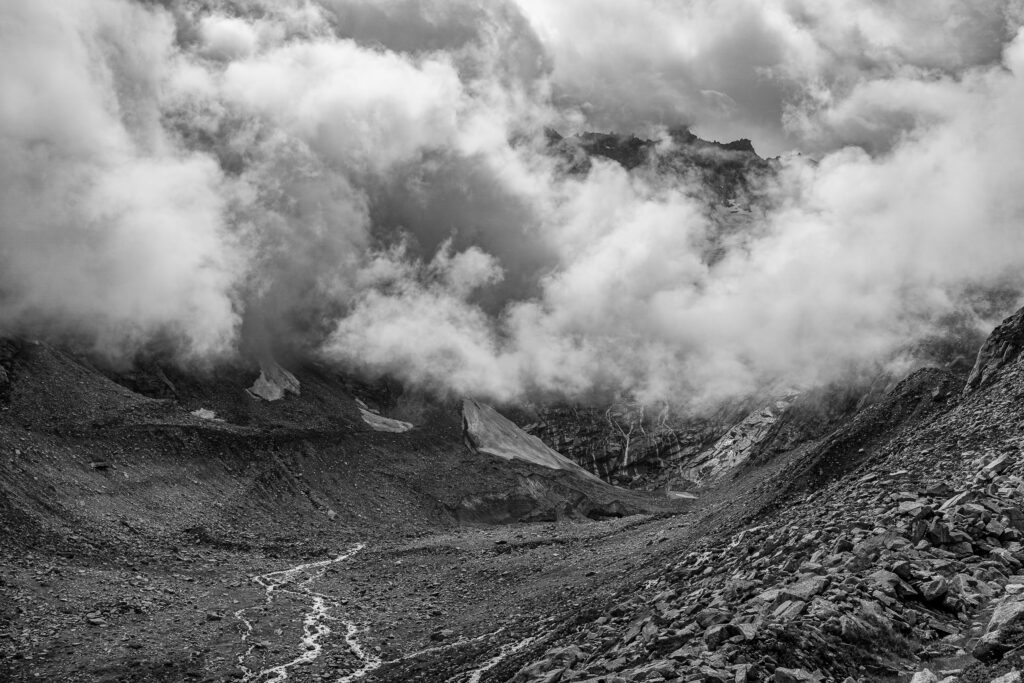 Schwarz-weiss Aufnahme von wolkenverhüllten Berggipfeln, Geröllhalde und Bergbach